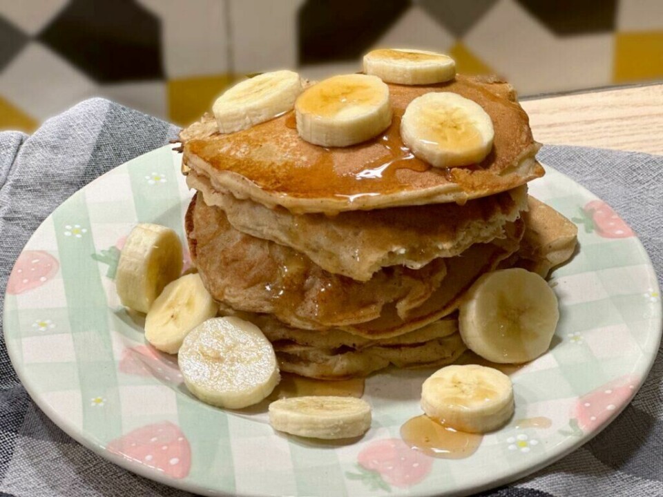 Nyt en sunn start på dagen med deilige bananpannekaker og ferske bananskiver! (Foto: Kreative Ideer/ Miriam Schjetlein) Bildet viser en fristende stabel av gyllenbrune bananpannekaker, toppet med skiver av fersk banan og drysset med honning eller lønnesirup. Pannekakene er tykke og luftige, servert på en dekorativ tallerken med et lyst mønster. Rundt stabelen ligger flere bananskiver som komplementerer retten, og alt dette står på en grå tekstil serviett.