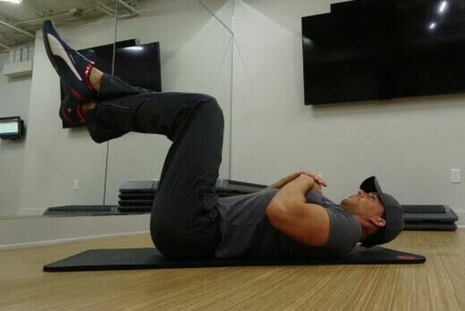 Crunches: Legg deg ned på gulvet, med føttene opp i været, i en 90-graders vinkel. (Foto: Cornelis Elander) Bilde av personlig trener Cornelis Elander som utfører en mageøvelse kjent som 'crunches' på en treningsmatte i et treningsrom. Han ligger på ryggen med knærne bøyd og føttene hevet i luften, krysset ved anklene. Hendene hans er krysset over brystet, og hodet og skuldrene er løftet fra bakken, noe som viser at han er i ferd med å kontrahere magemusklene. Mannen har på seg en mørk t-skjorte, mørke treningsbukser, en caps og treningssko. I bakgrunnen er det speilvegger, flere treningsmatter og en stor TV skjerm montert på veggen. Øvelsen han gjør fokuserer på å styrke magemusklene. Øvelsen fokuserer på å trene kjernemuskulaturen, og mage for en strammere, mage med synlige magemuskler og bedre holdning. Dette er en styrkeøvelse med fordeler for bein og rumpe, og en av styrkeøvelsene i vårt 30-dagers gratis treningsprogram og styrkeprogram.