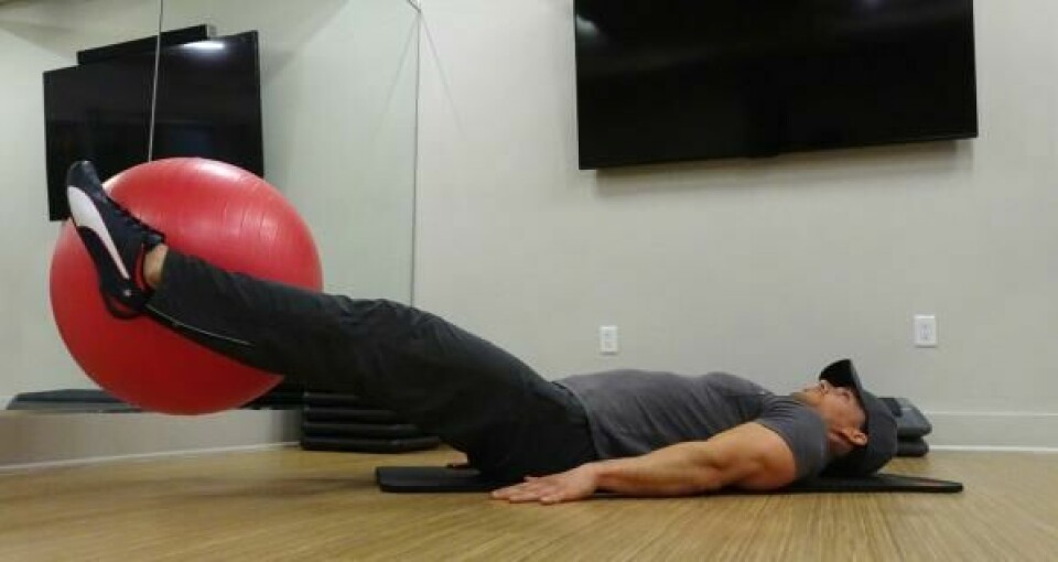 Liggende beinhev: Stram magen mens du løfter beina, strake oppover mot taket. (Foto: Cornelis Elander) Bilde av personlig trener Cornelis Elander som trener beinhev på treningsmatte og med en rød pilatesball. Han har på seg en grå t-skjorte, en svart treningsbukse og befinner seg i et treningsrom. Dette er en styrkeøvelse med fordeler for mage, core og bein, og er en av styrkeøvelsene i vårt 30-dagers gratis treningsprogram og styrkeprogram.