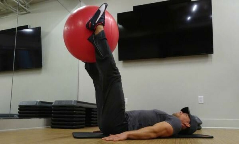 Liggende beinhev: Senk så beina rolig ned igjen til treningsmatta og utgangsposisjon. (Foto: Cornelis Elander) Bilde av personlig trener Cornelis Elander som trener beinhev på treningsmatte og med en rød pilatesball. Han har på seg en grå t-skjorte, en svart treningsbukse og befinner seg i et treningsrom. Dette er en styrkeøvelse med fordeler for mage, core og bein, og er en av styrkeøvelsene i vårt 30-dagers gratis treningsprogram og styrkeprogram.