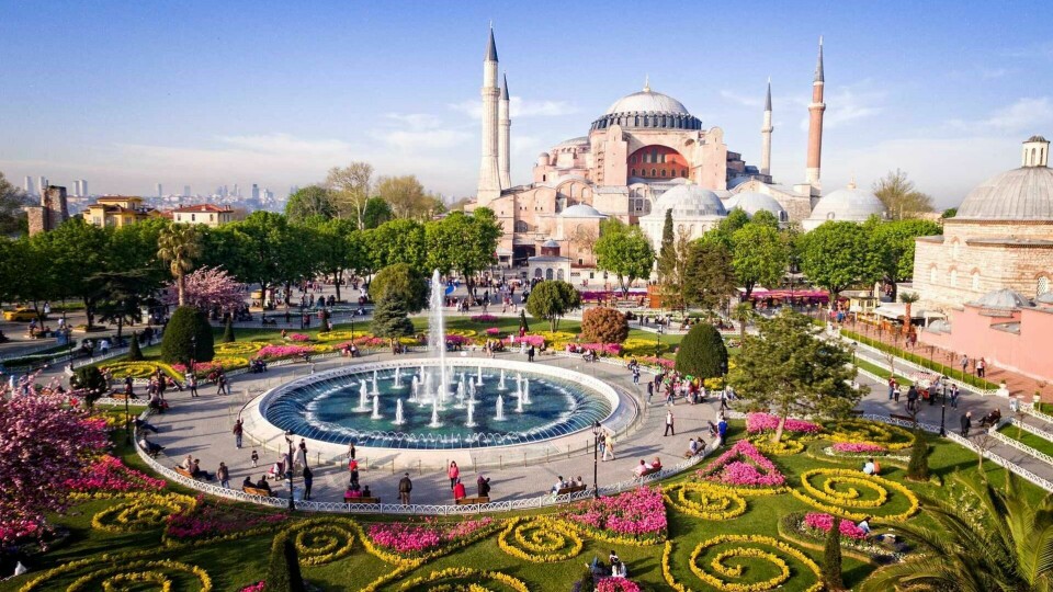 Istanbul byr på historiske skatter som Hagia Sophia og vakre parker. Et unikt reisemål der historie og moderne kultur møtes! (Foto: Apollo) Bildet viser den ikoniske Hagia Sophia i Istanbul, Tyrkia, med en stor, blomsterdekorert park og en fontene i forgrunnen. Hagia Sophia er en imponerende bygning med kupler og minareter, omgitt av frodige trær og fargerike blomsterbed som skaper en vakker og rolig atmosfære. Mange mennesker spaserer og nyter området i det solfylte været.