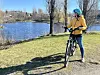 Bilde av dame på sykkel ute i solskinn i naturen. Hun har på seg en blå hjelm med solbriller og en gul dunjakke og hun står på en gressplen med Smestaddammen i bakgrunnen. Hun står ved en svart Buddy elsykkel til dame. Vi har snakket med sykkeleksperten som forteller hva du må tenke på når du skal kjøpe sykkel til voksne, enten det er til dame eller herre. Han forteller når det er lurt å gå for terrengsykkel, hybrid, bukkestyre, landeveissykkel, eller grussykkel (gravelsykkel) og hvilke sykler som passer til pendling.