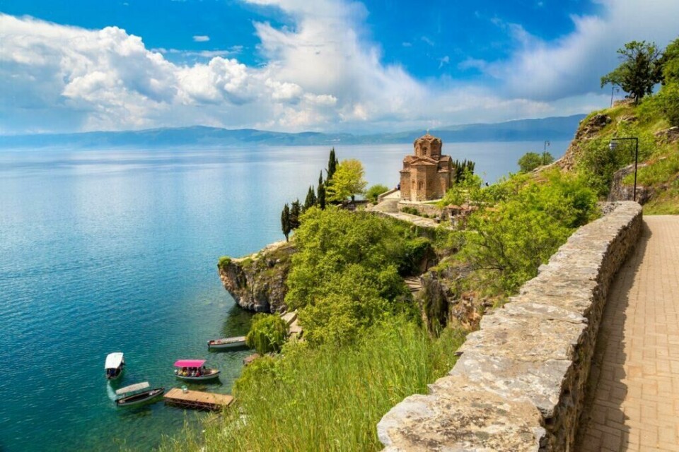 Nord-Makedonia er det billigste landet i Europa for nordmenn å feriere i. Landet byrblant annet på den spektakulære Ohridsjøen og den populære St John of Kaneo-kirken. (Foto: Colourbox) Bildet viser en pittoresk utsikt over Jovan Kaneo-kirken i Nord-Makedonia bygget på en klippe ved bredden av vakre Ohridsjøen. Kirken, som er laget av rødbrune steiner med detaljerte arkitektoniske utsmykninger, omgis av frodige grønne trær og busker. Foran kirken ligger en steinbelagt sti som fører langs klippens kant, og noen små båter er fortøyd i det klare blå vannet nedenfor. I bakgrunnen kan man se et fjellandskap og en dramatisk himmel med spredte skyer. Vi hjelper deg med å finne billig ferie, og svarer på spørsmål som hvor er det billig å reise, hva koster en halvliter øl, og avslører fem land som er kåret til de billigste i Europa for nordmenn å feriere i.