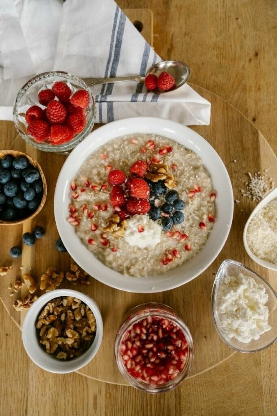 Havregrøt med cottage cheese, blåbær valnøtter og granateple. Dette er oppskriften på ernæringsfysiologens favorittfrokost! Bildet viser en frokostskål med havregrøt toppet med friske bær, nøtter og granateplefrø. Grøten er servert i en hvit bolle på et rundt trefjøl. Rundt grøten er det små skåler med ingredienser som blåbær, valnøtter, bringebær, granateplefrø, revet kokos og cottage cheese. En skje med bringebær hviler på en hvit og blåstripet kjøkkenklut. Bildet gir et inntrykk av en sunn og næringsrik frokost med ferske, naturlige ingredienser. Havregrøt med cottage cheese, blåbær valnøtter og granateple. Dette er oppskriften på ernæringsfysiologens favorittfrokost!