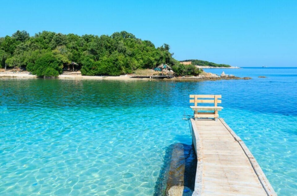 Albania byr på krystallklart hav blandet med spennende kultur og historie. Byen Ksamil er en av de mest populære destinasjonene i dette landet som også er billig å feriere i. (Foto: Colourbox) Bildet er det en idyllisk trebrygge som strekker seg ut i klart, turkisblått vann i Ksamil i Albania. På enden av bryggen er det en enkel trebenk vendt mot en liten, frodig øy med tett, grønn vegetasjon. Øya har en liten sandstrand og noen parasoller. I bakgrunnen kan man skimte en horisont av fjerne åser under en klar blå himmel. Vi hjelper deg med å finne billig ferie, og svarer på spørsmål som hvor er det billig å reise, hva koster en halvliter øl, og avslører fem land som er kåret til de billigste i Europa for nordmenn å feriere i.