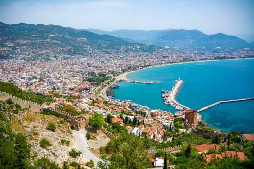 Tyrkia er en populær reisedestinasjon, og spesielt byene Antalya, Side og Alanya som du ser på bildet. Landet er også billig å feriere i, samt byr på både spennende historie og et yrende natteliv (Foto: Colourbox) Bildet viser en flott utsikt over Alanya i Tyrkia, med gamle steinmurer og det historiske vakttårnet, Det røde tårn, i forgrunnen. Byen er tett befolket med fargerike hus som strekker seg ned mot en lang sandstrand og en travel havn. Båter ligger forankret i det azurblå vannet, som strekker seg mot fjell i det fjerne under en klar blå himmel. Hele scenen er omringet av frodig grønne fjell og gir et inntrykk av en pulserende by som lever i harmoni med sin rike historie og naturskjønne omgivelser. Vi hjelper deg med å finne billig ferie, og svarer på spørsmål som hvor er det billig å reise, hva koster en halvliter øl, og avslører fem land som er kåret til de billigste i Europa for nordmenn å feriere i.