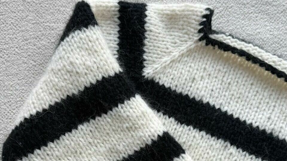 Nærbilde av Fridagenseren, en raglangenser fra Knitting by Emilie. (Foto: Knitting by Emilie) Nærbilde av en strikket genser med raglanfelling, der skulderpartiet viser de karakteristiske skrå linjene som dannes av fellingene. Genseren har et tidløst svart-hvitt stripemønster og er strikket i et jevnt og mykt garn. Raglankonstruksjonen gir en sømløs overgang mellom ermer og bol, noe som sikrer en behagelig passform og et stilrent uttrykk. Perfekt for både nybegynnere og erfarne strikkere som ønsker å lære mer om raglanfelling og raglanøkning