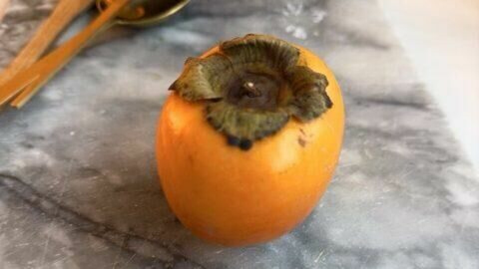 Kaki (persimmon) – en frisk og fargerik frukt som tilfører smak og farge til ethvert måltid. En hel kaki (persimmon) plassert på en marmorbordplate, omringet av gyldent bestikk og en morter i messing. Den sterke oransje fargen på frukten gir en vakker kontrast til bordplatens grå toner.