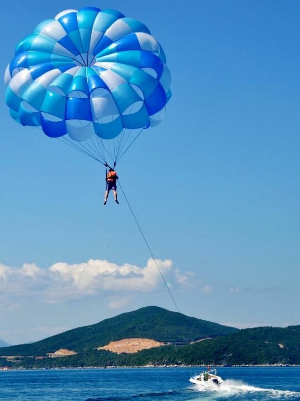 Vannski, bananbåt, vannscooter og en rekke andre vannaktiviteter er også populært på Gran Canaria. Bildet viser en person som driver med parasailing, hengende høyt oppe i luften under en stor blå og hvit fallskjerm. Personen er festet til en sele og blir dratt av en båt som kjører på vannet nedenfor. I bakgrunnen kan man se et landskap med grønne åser og fjell under en blå, skyfri himmel. Her får du ti ting du kan gjøre på Gran Canaria, Kanariøyene