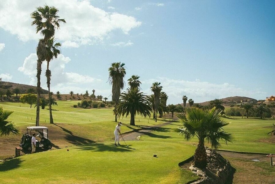 Hvis du liker golf kan du glede deg over at Gran Canaria også byr på flere golfbaner du kan sjekke ut. (Foto: Apollo) Bildet viser en golfbane med grønne fairways og palmer spredt rundt på området. I forgrunnen står en golfer som gjør seg klar til å slå en ball, mens en golfbil er parkert til venstre med en annen person ved siden av. Landskapet rundt golfbanen er kupert, og himmelen er blå med noen få skyer. Her får du ti ting du kan gjøre på Gran Canaria, Kanariøyene