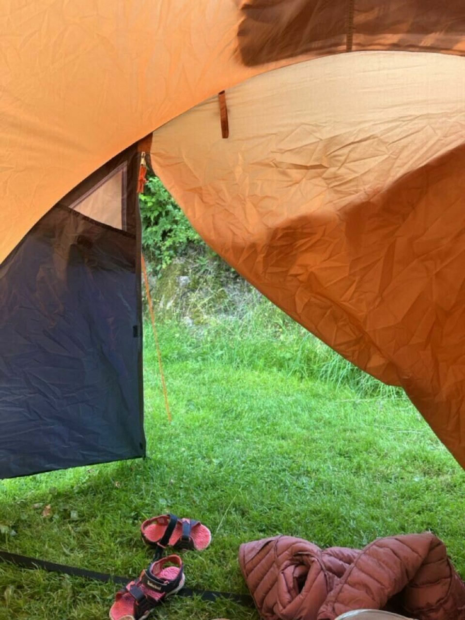 Telt-markedet er stort, og det finnes en rekke forskjellige telttyper å velge mellom. Enten du skal på telttur om sommeren eller vinteren, alene eller med familien, bilferie eller til fots, finnes det et telt som passer for deg. Utsikt fra innsiden av et telt, med teltduken i oransje og svart som rammer inn synsfeltet. Åpningen står delvis åpen mot en grønn gressplen og tett vegetasjon utenfor. På bakken foran teltåpningen ligger et par små, rosa og svarte sandaler, samt en sammenrullet, brun dunjakke. Lyset utenfra gir teltduken et varmt, gyllent skjær, og hele bildet gir en følelse av rolig morgenstemning eller en avslappet stund på telttur. Det finnes mange telt å velge mellom, blant annet campingtelt, helårstelt, pop-up telt, tarp og telt til bil. Vi har skrevet en guide som hjelper deg å finne riktig telt til teltturen.