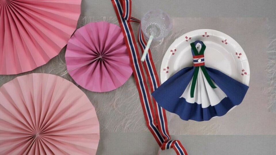 Hvis du bytter ut farger på servietter og bånd, kan du brette bunad-serviett som ligner din egen bunad. (Foto: Kreative Ideer/ Miriam Schjetlein) Pyntet festbord med 17.mai-bånd, rosa vifter og grå duk. Papptallerken med bunad i blått og hvitt brettet av serviett.