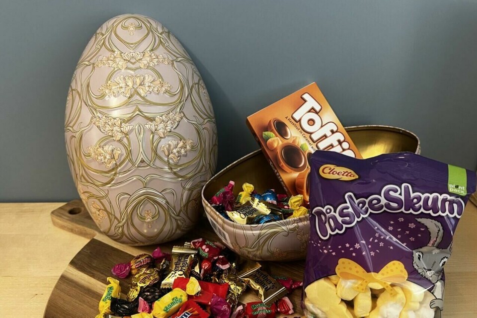 Testvinner: Et vakkert påskeegg med elegant mønster, fylt med sjokolade, karameller og mykt påskeskum. Perfekt for påskekosen! På bildet ser vi et stort, vakkert påskeegg med en elegant, gammeldags mønstring i duse farger, hovedsakelig beige og gull. Påskeegget er delt i to, hvor den ene halvdelen ligger åpen og avslører et fylt påskeegg med en rik blanding av godteri. Det bugner av sjokolade, karameller og andre fargerike innpakkede søtsaker. Blant godsakene ligger også en eske Toffifee og en pose Cloetta Påskeskum med myke, gule og hvite skumgodterier. Alt dette ligger på et rundt trefat mot en blågrå vegg i bakgrunnen, noe som gir en koselig og stilfull påskestemning.