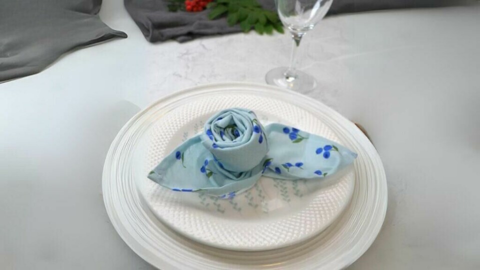 Forvandle borddekkingen med en vakker rosebrettet serviett – en enkel og elegant detalj! (Foto: Kreative Ideer/ Miriam Schjetlein) En lyseblå tøyserviett med et mønster av små blå blomster er brettet til en elegant roseform, plassert midt på en hvit tallerken med diskret mønster. Servietten er kunstferdig brettet slik at rosen står ut i midten, med endene formet som blader på hver side. Tallerkenen står på en lys marmorbakgrunn, og i bakgrunnen skimtes et vannglass og grønne dekorative elementer.