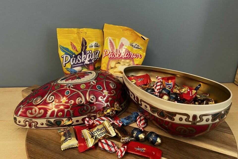 Fabergé-påskeegg fylt med sjokolade, karameller og påskeskum – en luksuriøs testvinner til påskekosen! 🐣✨ På bildet ser vi et eksklusivt Fabergé-inspirert påskeegg med en dyp rød bunnfarge, dekorert med detaljer i gull, blått og hvitt, som gir det et luksuriøst og kongelig preg. Egget er delt i to, hvor den ene halvdelen ligger åpen og avslører et fylt påskeegg med en blanding av sjokolade og karameller, blant annet Werther’s Original og Daim. Rundt egget ligger flere sjokoladebiter løst, og bak det står to poser med påskegodteri – én med dragébelagte sjokoladeegg og én med myke påskeskum-godterier. Bakgrunnen er en blågrå vegg, og alt er plassert på et rundt trefat, noe som gir en eksklusiv, men samtidig koselig påskestemning.