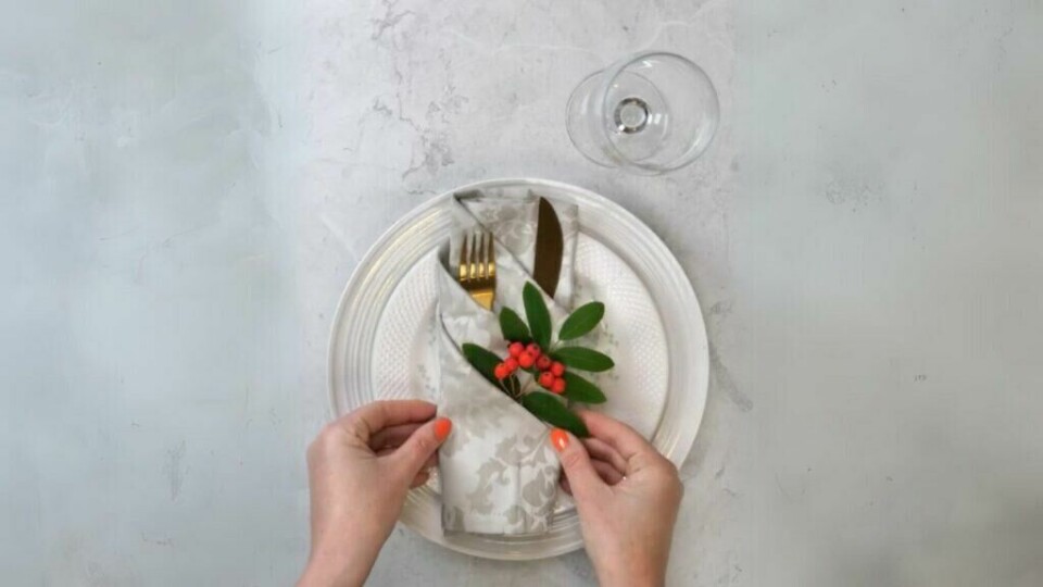 Lær å brette en elegant serviett med bestikklomme. (Foto: Kreative Ideer/ Miriam Schjetlein) En enkel guide til å brette en tøyserviett med bestikklomme. På bildet ser vi en hvit tallerken mot en lys marmorbakgrunn, der en mønstret serviett er brettet slik at den danner en lomme til gullfarget bestikk. Servietten er pyntet med en grønn kvist og røde bær for et dekorativt preg. Personen med oransje neglelakk tilpasser servietten forsiktig, og viser hvordan man kan organisere borddekkingen med stil.