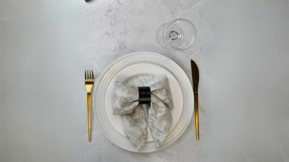 Pynt bordet med en elegant sløyfebrettet serviett for et stilfullt uttrykk. (Foto: Kreative Ideer/ Miriam Schjetlein) Et sofistikert bordoppsett med en hvit tallerken på en lys marmorbakgrunn. Midt på tallerkenen er en dekorativ serviett brettet som en sløyfe, festet med en svart serviettring. Servietten har et mønster i lysegrått, som gir et klassisk og elegant preg. Gullfarget bestikk er plassert symmetrisk på hver side av tallerkenen, og et tomt vinglass står øverst til høyre, klart til bruk.