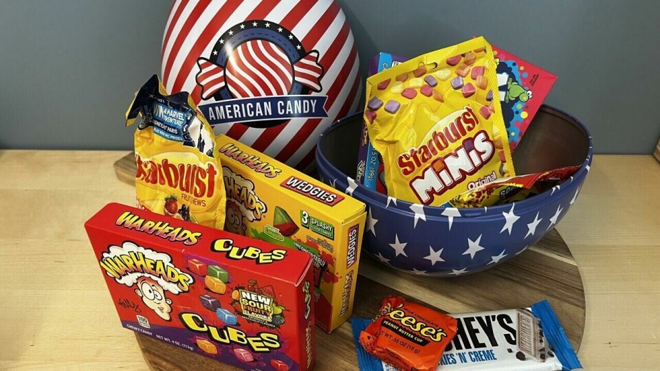 Test av påskeegg 2025: Her er årets vinner På bildet ser vi et påskeegg med amerikansk godteri, designet med et patriotisk amerikansk tema. Egget er rødt og hvitt stripet med en stor logo som sier 'American Candy' og har et emblem med stjerner og striper. Den nederste delen av egget er blå med hvite stjerner, inspirert av det amerikanske flagget. Egget er delt i to, og innholdet er en spennende blanding av klassisk amerikansk godteri. Blant godteriet finner vi kjente merker som Warheads Cubes (sure godteribiter), Reese’s Peanut Butter Cups (sjokolade med peanøttsmør), Starburst Minis (fruktkarameller), Hershey’s Cookies 'n' Creme (hvit sjokolade med sjokoladebiter) og andre fargerike godteriposer. Godteriet ligger både inne i egget og på et rundt trefat foran det, med flere poser og sjokoladebiter pent arrangert. Bakgrunnen er en blågrå vegg som gir et rolig bakteppe til de sterke fargene på egget og godteriet.