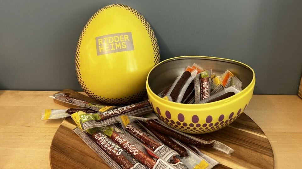 Påskeegg fra Ridderheims fylt med smakfulle ølpølser i ulike varianter. Perfekt for deg som foretrekker salt snacks fremfor søtsaker! (Foto: Ole Kristian Aaknes) På bildet ser vi et påskeegg fra Ridderheims, som skiller seg ut fra tradisjonelle påskeegg med godteri. Egget er gult med lilla prikker langs kanten og har en metallisk innside. På forsiden av egget står det 'Ridderheims' i store gule bokstaver på en lilla bakgrunn. Egget er delt i to, og den nederste halvdelen er fylt med individuelt pakkede ølpølser i ulike smaker. Noen av pølsene ligger også spredt utover et rundt trefat foran egget. Vi ser etiketter med navn som 'Cheezes' (med ostesmak) og 'Hot Amigo' (med krydder). Bakgrunnen er en blågrå vegg, som gir en rolig kontrast til det sterke gule egget.