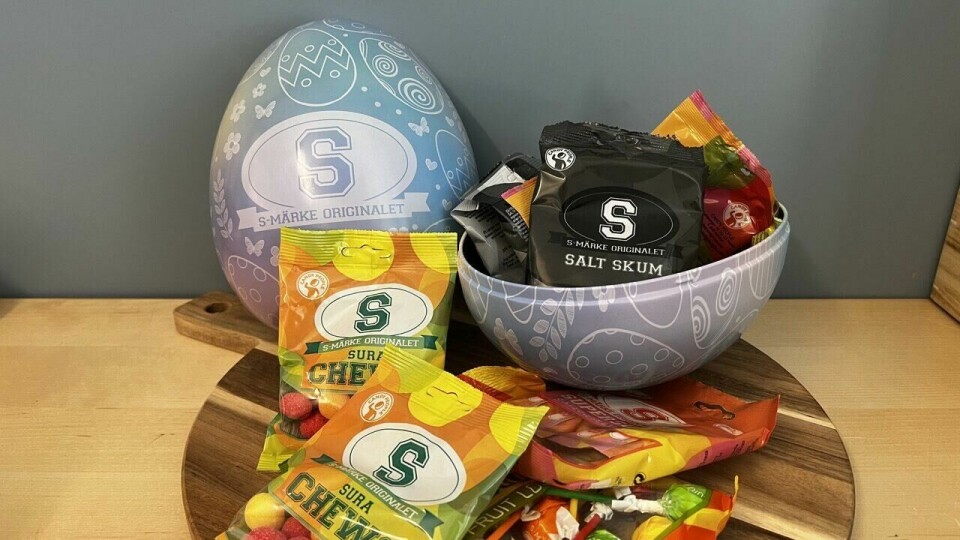 S-merke påskeegg fylt med sure og salte godterifavoritter som 'Sura Chews' og 'Salt Skum' – perfekt for deg som elsker en syrlig påske! På bildet ser vi et S-merke påskeegg, som er laget for dem som elsker sure og salte smaker. Egget er lyseblått med hvite mønstre, inkludert påskemotiver som egg, blomster og virvler. På forsiden av egget står en stor 'S' med teksten 'S-Märke Originalet' under.Egget er delt i to, og den nederste halvdelen er fylt med poser av S-merke-godteri. Blant innholdet ser vi poser med 'Salt Skum' (i svart innpakning), 'Sura Chews' (i grønn og gul innpakning) og fruktkarameller. Flere poser ligger spredt på et rundt trefat foran egget, noe som gir et innbydende uttrykk. Bakgrunnen er en blågrå vegg, som skaper en rolig kontrast til de fargerike godteposene.
