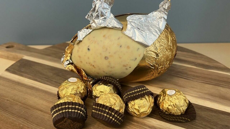 Ferrero Rocher påskeegg i hvit sjokolade med sprø hasselnøttbiter, delvis pakket ut fra sin luksuriøse gullfolie. Omkranset av klassiske Ferrero Rocher-praliner – en eksklusiv påskegodbit! (Foto: Ole Kristian Aaknes) På bildet ser vi et Ferrero Rocher påskeegg i hvit sjokolade. Egget er delvis pakket ut fra sin gylne folieinnpakning, som er krøllet tilbake for å avdekke den kremete, hvite sjokoladen. Sjokoladen har en sprø tekstur med synlige biter av ristede hasselnøtter.Foran egget ligger flere Ferrero Rocher-praliner, innpakket i sin ikoniske gullfolie med små, brune papirformer. Egget og sjokoladene er plassert på et rundt trefat, og bakgrunnen er en blågrå vegg som skaper en nøytral kontrast til de varme gulltonene.