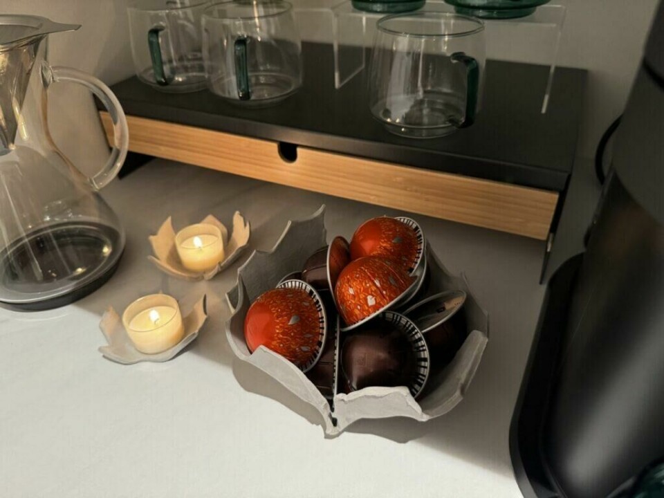 En leirskål formet som et blad fylt med kaffekapsler, plassert i et koselig kaffehjørne. (Foto: Kreative ideer/ Aurora Lange) Bildet viser en håndlaget leirskål formet som et blad, fylt med kaffekapsler i ulike farger. Skålen står på en lys overflate, og ved siden av er to mindre leirskåler, også formet som blader, som fungerer som telysholdere med tente lys. I bakgrunnen er det en glasskaraffel, gjennomsiktige kaffekopper med grønne hanker, og en liten hylle som utgjør en del av kaffehjørnet. Hele oppsettet skaper en varm og innbydende atmosfære, med de håndlagde leirskålene som gir et unikt og dekorativt innslag.
