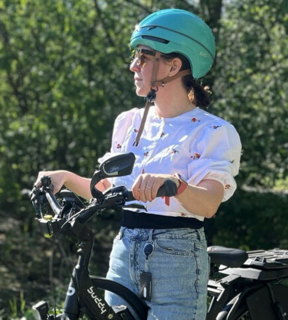 Miriam Schjetlein, vår egen el-syklist, gir sine beste tips til genialt utstyr du bør vurdere om du har elsykkel! Bilde av dame som står med en elsykkel til dame fra Buddy Bike. Hun har på seg en hvit bluse, dongeribukser og en blå sykkelhjelm og står med elsykkelen sin på en gressplen med et vann i bakgrunnen i sol og sommervær. Vi gir 20 geniale tips til utstyr til elsykkel, og alt fra sykkelvesker, mobilholder, forsikring til elsykkel, sykkelhjelm som er best i test til elsykkel og best i test sykkellås.