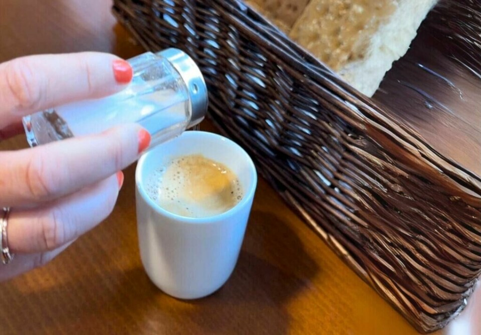 En uventet smaksvri – litt salt i kaffen kan overraske smaksløkene. (Foto: Kreative Ideer/ Miriam Schjetlein) En nærbilde viser en hånd med røde negler som drysser salt fra en saltbøsse ned i en kopp med kaffe. Kaffen har et lett skumlag på toppen. Ved siden av står en kurv med brød, og overflaten er et mørkt trebord som gir en varm atmosfære.
