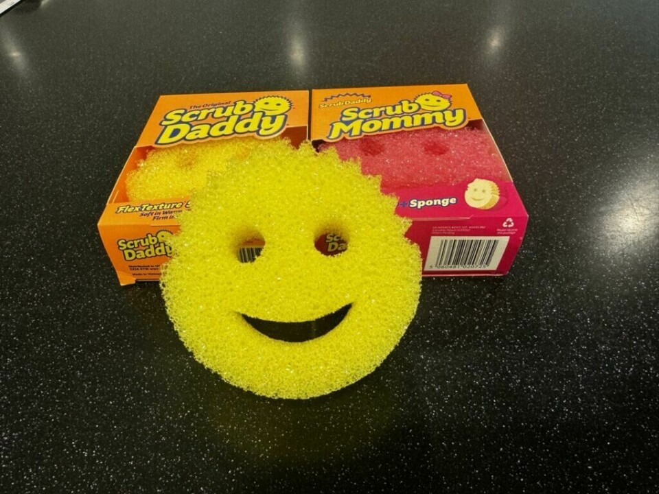 En gul Scrub Daddy-svamp smiler foran sine originale emballasjer for både Scrub Daddy og Scrub Mommy. Bildet viser en gul, smilende skuresvamp (Scrub Daddy) plassert foran to esker: en oransje Scrub Daddy-eske og en rosa Scrub Mommy-eske. Den gule svampen har et ansikt med øyne og en smilende munn, og den står på en mørk overflate med små hvite prikker. De fargerike eskene i bakgrunnen har logoene og bilder av svampene, som matcher svampens design og farger.