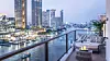 Bildet viser luksuriøse Mandarin Oriental, Bangkok, plassert ved Chao Phraya-elvens bredd i Thailand. Hotellets elegante, historiske bygninger er omgitt av frodig grøntområder og tropiske hager, med en stor, glitrende elv som bakteppe. På terrassen kan man se hvite parasoller og stilige gjester som nyter utsikten. Lyset fra solnedgangen speiler seg i elven, og atmosfæren utstråler eleganse og ro.
