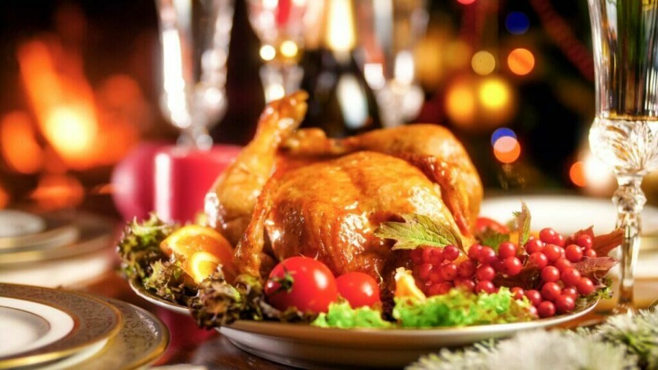 Saftig, ovnsbakt kalkun – en imponerende hovedrett til julemiddagen, komplett med smakfullt tilbehør og juledekor. Et gyllent, ovnsstekt kalkun på et dekorativt fat omgitt av friske salatblader, kirsebærtomater, appelsinskiver og røde bær. I bakgrunnen skimtes et koselig juleoppsett med tente lys og glitrende bokeh-lys, som gir en varm og festlig atmosfære. Kalkunen har en glinsende, perfekt stekt overflate.