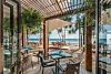 Bildet viser en eksklusiv strandrestaurant på Iniala Beach House i Thailand. Utsikten åpner seg mot en gyllen sandstrand og det asurblå havet, mens bordene er elegant dekket med blå servietter og vannflasker. Restauranten har en moderne, åpen design med trekonstruksjoner og hengende grønne planter, som skaper en harmonisk atmosfære. Mykt sollys strømmer gjennom pergolataket og kaster skygger på tregulvet, mens havbrisen beveger de lette solseilene over utendørs spiseområdet.