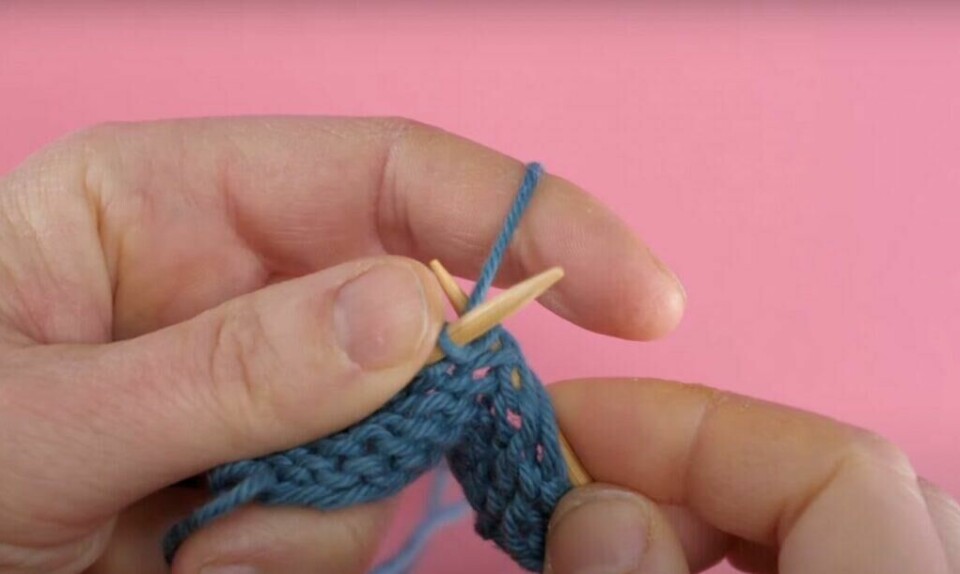 2) Vri høyre strikkepinne tilbake hent garnet. (Foto: ABC Brand Studio) Bilde av hvordan strikke vrange masker med blått garn, strikkepinner i tre og rosa bakgrunn