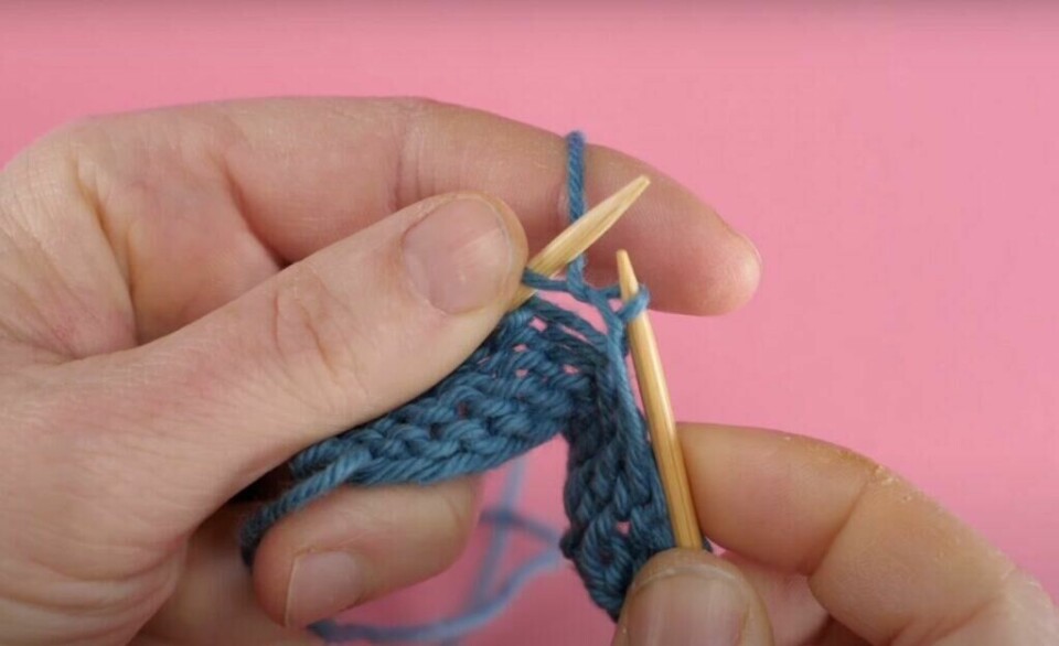4) Dra garnet med deg tilbake og gjennom masken. Slipp den gamle masken av venstre pinne. (Foto: ABC Brand Studio) Bilde av hvordan strikke vrang med blått garn, strikkepinner i tre og rosa bakgrunn