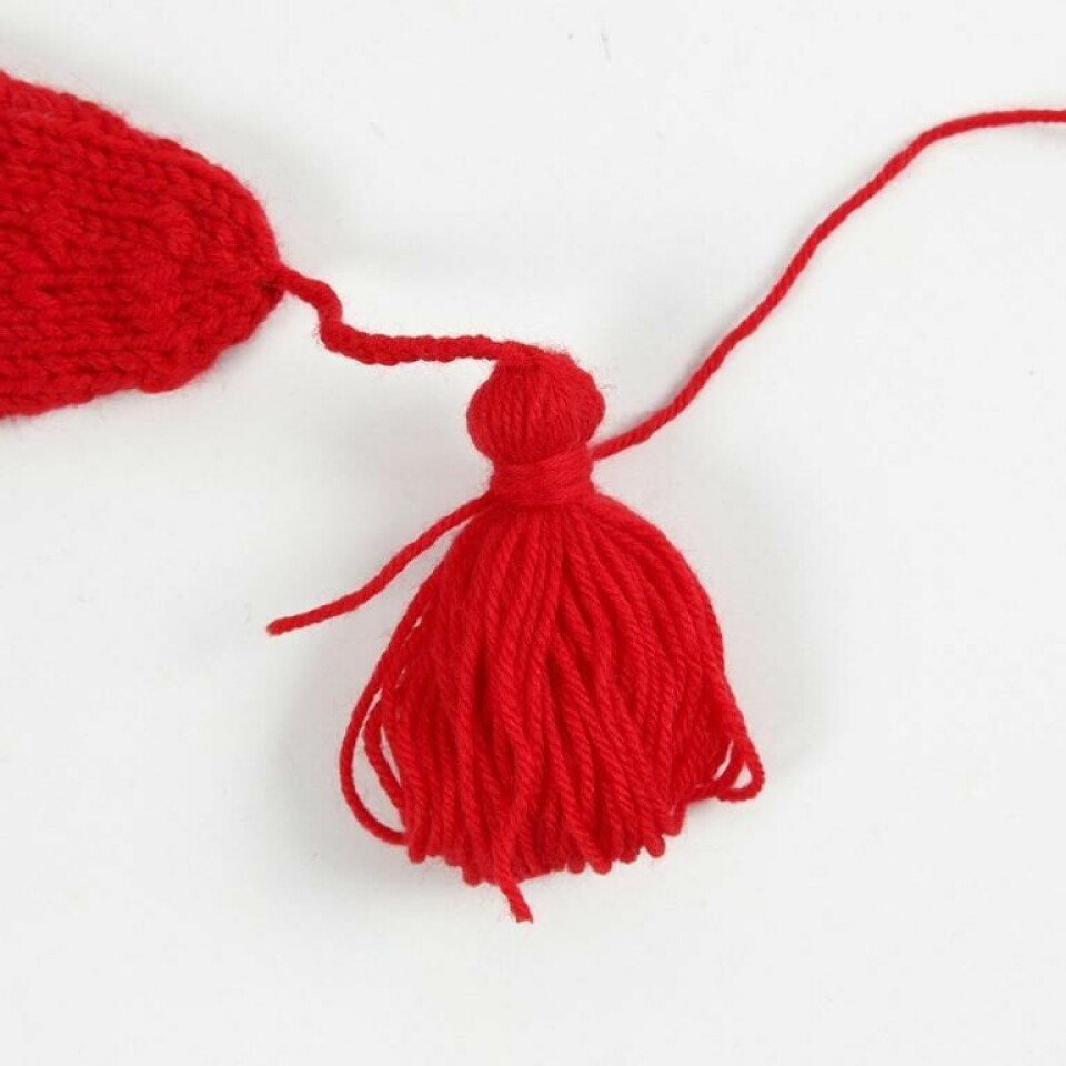 Steg 4. Fjern pappen og knyt enden av en lang garntråd rundt halvdelen. Vri garnet rundt mange ganger og bind to knuter. Klipp av i samme lengde som dusken. (Foto: CC hobby) Bilde av rød tråd surret rundt rød dusk til strikkeoppskrift nisselue