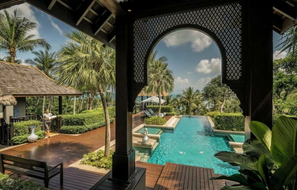 Resortet, Four Seasons Resort på Koh Samui, kjent fra den nye sesongen av The White Lotus, ligger på en idyllisk Thailandsk øy. Her finner du elegante villaer, luksuriøse fasiliteter og en atmosfære av fredelig luksus. (Foto: Hotels.com) Bildet viser en luksuriøs resort med tropisk beliggenhet, omgitt av palmer og frodig grønn vegetasjon. Perspektivet er fra en overbygd terrasse med mørkt treverk og et dekorativt buet vindu med et mønstret gitter, som rammer inn utsikten. Foran oss strekker et stort, glitrende evighetsbasseng seg utover mot horisonten, med et elegant oppbygget design og små fossefall. På venstre side av bildet står en person i lette klær, sannsynligvis en hotellansatt eller en gjest, ved en liten bungalow med stråtak. En gangvei i mørkt treverk fører gjennom resortens velstelte hageområder. I bakgrunnen skimtes det glitrende blå havet mellom palmene, under en himmel med hvite skyer. Atmosfæren utstråler eksklusiv ro og velvære, perfekt for en luksuriøs ferieopplevelse.