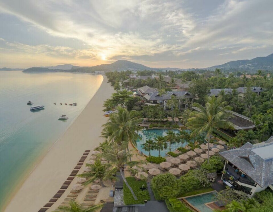 Luftfoto av den luksuriøse resorten Anantara Bophut på Koh Samui, en av innspillingslokasjonene for den nye sesongen av The White Lotus. Her møtes hvite sandstrender, tropiske hager og elegante bassenger i en perfekt kombinasjon av luksus og eksotisk ro. En destinasjon like dramatisk og vakker som serien selv. (Foto: Hotels.com) Bildet viser en spektakulær luksusresort ved en rolig sandstrand i Thailand, fanget i et mykt gyllent lys fra en solnedgang i horisonten. Utsikten er fra luften og gir et vidstrakt perspektiv over resortens frodige hageområder, svømmebassenger og havet som strekker seg ut mot horisonten. I forgrunnen ligger et stort basseng omgitt av solsenger og parasoller, med palmetrær spredt utover, noe som gir stedet en tropisk, eksklusiv atmosfære. Ved bassenget og i hagen er det luksuriøse bygninger med mørke tretak og åpen arkitektur, harmonisk integrert i naturen. Den lange, gylne sandstranden strekker seg langs kysten, med rader av solsenger og parasoller plassert pent i rekke. Små båter ligger forankret rett utenfor kysten, klare for eventyr på havet. Bak resorten brer grønnkledde åser seg utover, med små landsbyer og hus spredt mellom palmene. Stemningen er rolig og idyllisk, med et perfekt møte mellom luksus og naturlig skjønnhet.