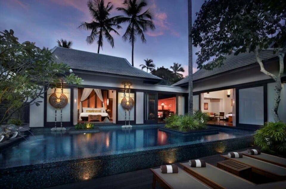 Anantara Mai Khao Villas i Phuket, en av innspillingslokasjonene for The White Lotus sesong 3. Med sitt spektakulære infinity-basseng, elegante Thailandske design og eksklusive omgivelser, gir denne resorten en perfekt ramme for dramatikken og dekadensen i serien. (Foto: Hotels.com) Bildet viser en luksuriøs privat villa i et eksklusivt feriested, omgitt av tropisk natur i skumringens myke lys. Foran villaen ligger et stort, mørkeblått infinity-basseng, hvor vannet speiler den vakre himmelen som skifter i lilla og rosa toner i solnedgangen. Villaen har et tradisjonelt, men moderne thailandsk design med lave tak, mørke tretak og store glassdører som åpner opp til de elegante rommene innenfor. Midt i bildet ser vi en romantisk himmelseng med hvite gardiner, vakkert belyst og omringet av sofistikerte dekorative elementer. På hver side av inngangen henger store, kunstneriske lyktelamper med thailandske mønstre. Til høyre for sengen skimtes en innendørs stue med varme lys, komfortable møbler og en rolig atmosfære. På venstre side av bildet fører bassengkanten mot en tropisk hage med palmetrær og grønne busker, som tilfører en følelse av ro og eksklusivitet. I forgrunnen ligger flere elegante solsenger med pent rullede håndklær, klare for gjester som ønsker å slappe av i denne luksuriøse tilbaketrekningen. Stemningen er en perfekt blanding av eksklusivitet, privatliv og tropisk harmoni.