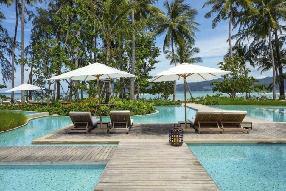 Det luksuriøse bassengområdet på Rosewood Phuket, en av innspillingslokasjonene for The White Lotus sesong 3. Her finner du blant annet restauranten hvor noe av innspillingen foregår i serien. (Foto: Hotels.com) Bildet viser en luksuriøs tropisk resort med et elegant bassengområde, omgitt av frodig grønn vegetasjon og høye palmetrær. En bred trebro leder over et krystallklart, lysblått basseng og ender ved to solsenger med hvite puter, plassert under store, hvite parasoller. Bak solsengene skimtes et større svømmebasseng med avrundede kanter, designet for å smelte naturlig inn i omgivelsene. Rundt bassenget vokser grønne planter og blomster, mens palmetrærne gir en eksotisk følelse av paradis. I bakgrunnen åpner landskapet seg mot en idyllisk sandstrand og et turkist hav. Små bølger ruller inn mot kysten, og fjellene i horisonten danner en vakker bakgrunn. Over det hele svever en skyfri, lys blå himmel, som understreker den avslappende og luksuriøse stemningen. Stemningen er rolig og eksklusiv – et sted hvor man kan nyte solen, slappe av og ta inn den spektakulære utsikten over havet.