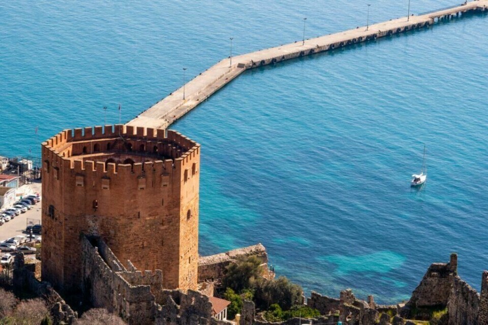 Besøker du Alanya i Tyrkia kan vi anbefale å ta turen til det røde tårnet, Kizil Kule! (Foto: Colourbox) Bilde av det røde tårnet i Alanya, Tyrkia. I bakgrunnen ser du blått hav og en molo. Her finner du fem ting du må se og gjøre langs Tyrkias kyst, som blant annet besøke Alanya og Side. Du finner også billige reiser her.