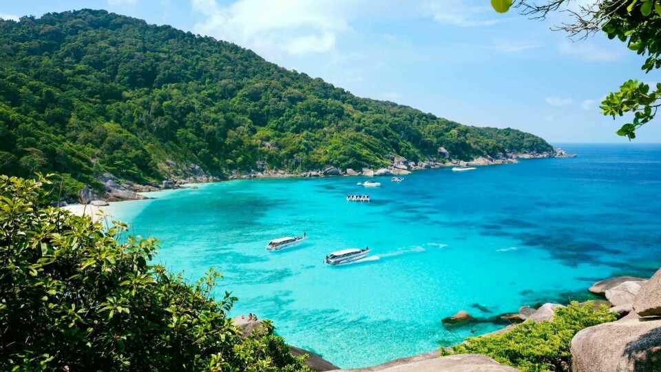 Et populært reisemål om vinteren er Thailand. (Foto: Apollo) Dette bildet viser en nydelig bukt i Thailand, et ideelt reisemål for en vinterreise til sol og varme. Bukten er fylt med krystallklart, turkisblått vann som glitrer under solen. I bakgrunnen strekker en frodig, grønnkledd åsside seg opp fra kysten, med tett, tropisk vegetasjon som dekker hele området. Flere små båter ligger forankret i bukten, klare til å ta besøkende på eventyr, kanskje til snorkling eller utforsking av de nærliggende strendene. Bildet oser av eksotisk skjønnhet og fred, med klare, solrike himler som gir en følelse av å være langt unna vinterens kulde. Dette gjør Thailand til en perfekt flukt fra den kalde vinteren, hvor man kan nyte tropiske omgivelser og varme temperaturer.