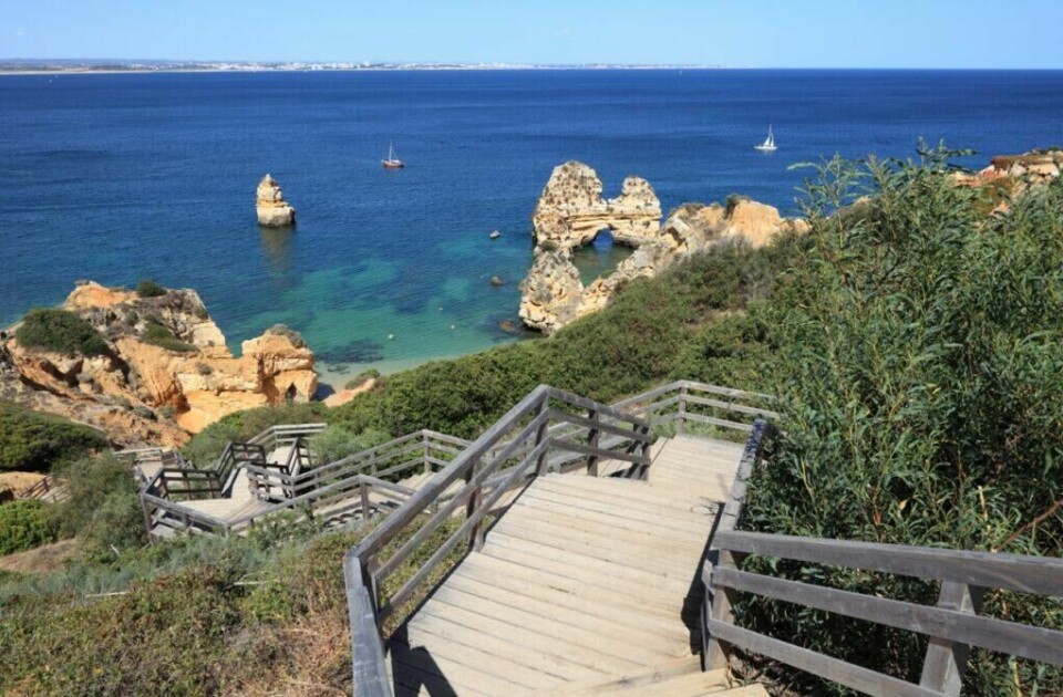 Lagos byr på vakre strender og blått hav langs Algarvekysten i Portugal. (Foto: Colourbox) Bildet viser en naturskjønn utsikt over Lagos-kysten i Algarve, Portugal. En tretrapp med rekkverk slynger seg nedover en grønnkledd klippe mot en liten strand og det krystallklare, blågrønne vannet. I bakgrunnen stiger steinformasjoner dramatisk opp fra havet, og to seilbåter flyter rolig på vannet. Det klare været gir en vidstrakt utsikt over havet og kystlinjen i det fjerne. Vi hjelper deg med å planlegge ferien til Algarve-kysten i Portugal. Vi gir deg alt fra tips til byer; Lagos, Faro, Tavira, Albufeira og Sagres. Samt hvor du kan finne hotell langs Algarve-kysten og shoppingmulighetene.