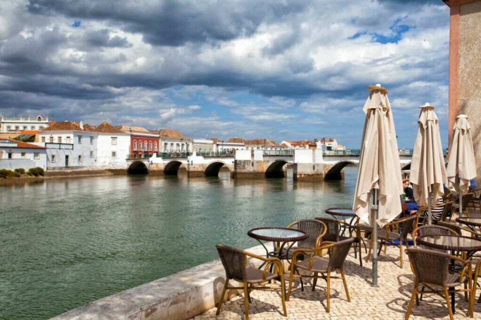 Byen Tavira i Portugal er kjent for sine mange broer og kalles ofte for Algarvekystens Venezia. (Foto: Colourbox) Bildet viser byen Tavira langs Algarve-kysten i Portugal. I forgrunnen er det et utendørs kaféområde med tomme bord og stoler, samt sammenfoldede parasoller. I bakgrunnen ser vi en gammel steinbro med flere buer som strekker seg over en rolig elv. På den andre siden av elven står sjarmerende, fargerike bygninger med tradisjonell portugisisk arkitektur under en dramatisk, skyet himmel. Vi hjelper deg med å planlegge ferien til Algarve-kysten i Portugal. Vi gir deg alt fra tips til byer; Lagos, Faro, Tavira, Albufeira og Sagres. Samt hvor du kan finne hotell langs Algarve-kysten og shoppingmulighetene.