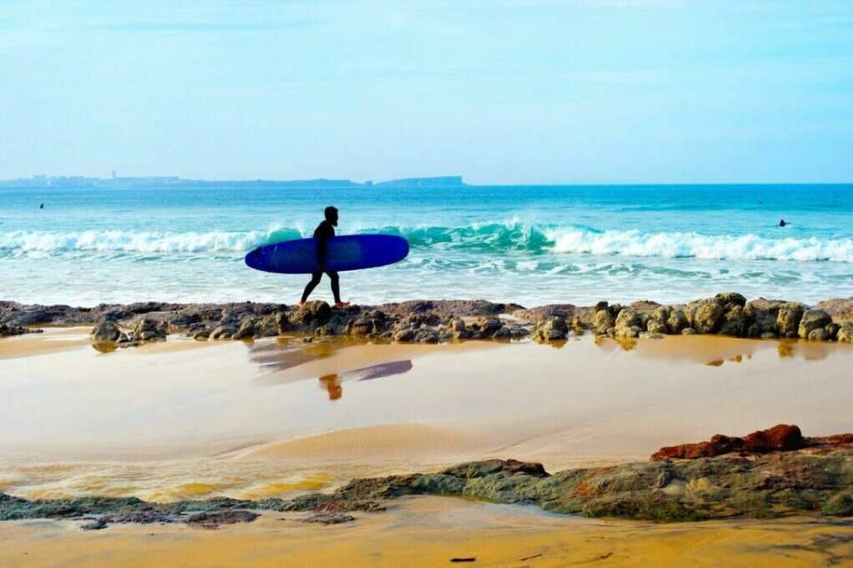 Liker du å surfe er byen Sagres langs Algarvekysten i Portugal helt klart destinasjonen for deg! (Foto: Colourbox) Bildet viser en surfer som går på en strand i Sagres, langs Algarve-kysten i Portugal. Surferen bærer et blått surfebrett under armen og er kledd i en våtdrakt. I bakgrunnen ruller bølgene inn mot kysten, og den klare, blå himmelen møter horisonten. Man kan også skimte en annen surfer i vannet. Vi hjelper deg med å planlegge ferien til Algarve-kysten i Portugal. Vi gir deg alt fra tips til byer; Lagos, Faro, Tavira, Albufeira og Sagres. Samt hvor du kan finne hotell langs Algarve-kysten og shoppingmulighetene.