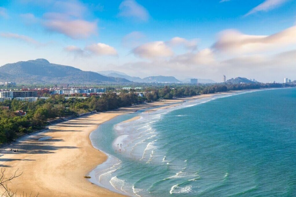 Hua Hin i Thailand er en av landets eldste badebyer og er kjent for sin avslappede atmosfære og som et populært feriested for den thailandske kongefamilien i over hundre år. (Foto: Colourbox) En lang, bred sandstrand med bølger som ruller inn mot land i Hua Hin, Thailand. Strandkanten er omgitt av frodig vegetasjon og bygninger i bakgrunnen, med åser og fjell som stiger opp i det fjerne under en blå himmel med noen lette skyer. hjelper deg med å finne reise til Thailand, enten du vil ha billige reiser med restplasser, eller kanskje pakkereiser til de populære destinasjonene Phuket, Hua Hin og Krabi. Her finner du også hotell i Thailand.