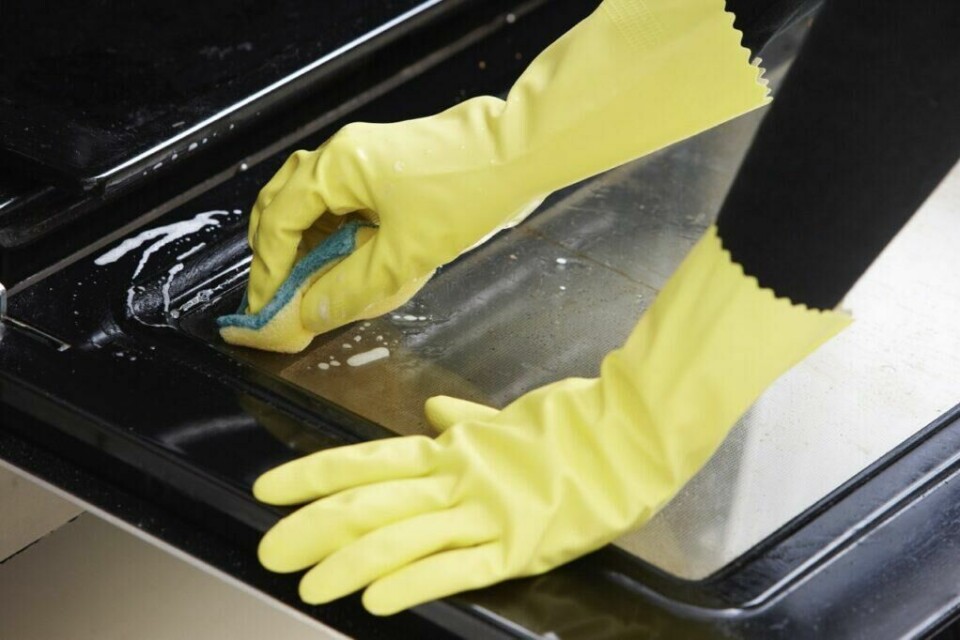 Det må gjerne både skrubbing og skuring til når stekeovnen skal rengjøres, men med dette trikset blir vaskejobben lekende lett! (Foto: Colourbox) Bilde av hender med vaskehansker som vasker døren på en stekeovn med en gul svamp.