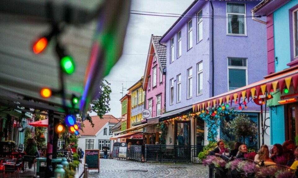 Øvre Holmegate er gaten i Stavanger sentrum der alle husene i østre del av gaten (mellom Kirkegata og Østervåg) er malt i flotte farger. Her finner du både kafeer, nisjebutikker og kunstutsalg. (Foto: Martin Håndlykken / Visit Norway) Folk på kafé i fargegaten i Stavanger. Full av fargerike trehus, gul, blå, lilla, rosa, grønn og oransje, med lyslenke i front.