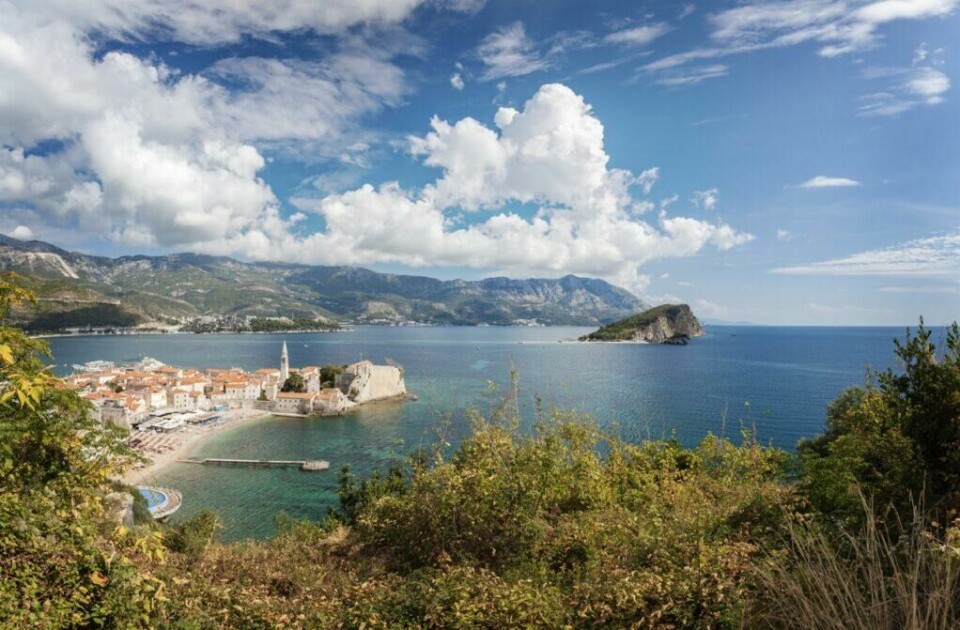 Budva i Montenegro er en populær destinasjon for de som søker vakre strender og krystallklart hav langs Middelhavet. (Foto: Colourbox) utsikt over Budva, Montenegro, sett fra en høyde. Byen ligger ved kysten og er omkranset av høye fjell i bakgrunnen. Gamlebyen med sine lyse bygninger og et prominent kirketårn ligger ved en liten halvøy, flankert av en stor strand og det krystallklare Adriaterhavet. Montenegro er et spennende land langs Middelhavet som er rangert til å være et billig land for nordmenn å feriere i. Vi hjelper deg med å finne pakkereiser som inkluderer fly og hotell, samt restplasser til Montenegro.