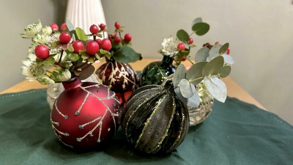kap julestemning med en krans av julekuler fylt med naturens vakre detaljer. (Foto: Kreative Ideer/ Miriam Schjetlein) Bildet viser en kreativ og stemningsfull borddekorasjon der julekulene er arrangert i en kransform. Kulene, i røde, grønne og sorte nyanser med glitrende dekorasjoner, fungerer som små vaser fylt med vintergrønt, røde bær og eukalyptusblader. Kransen er plassert på en mørkegrønn duk, noe som fremhever julestemningen og skaper et elegant uttrykk. Detaljene i både kulenes mønster og de naturlige elementene kommer tydelig frem i nærbildet.