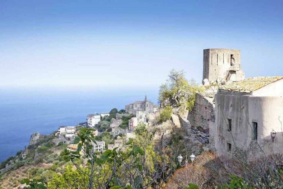 Sicilia er det som kan ligne en fotball ved 'tåen' på Italia på kartet. Øya er middelhavet største og et populært reisemål. (Foto: Apollo) Borg ogbebyggelse ned en ås på Sicilia, med hav og blå himmel i bakgrunn.